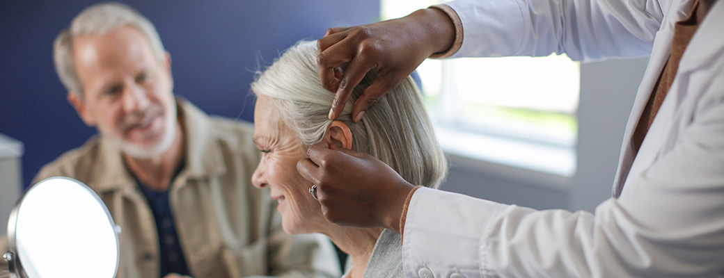 Image of woman being fitted for a hearing aid by a hearing care professional as her spouse watches